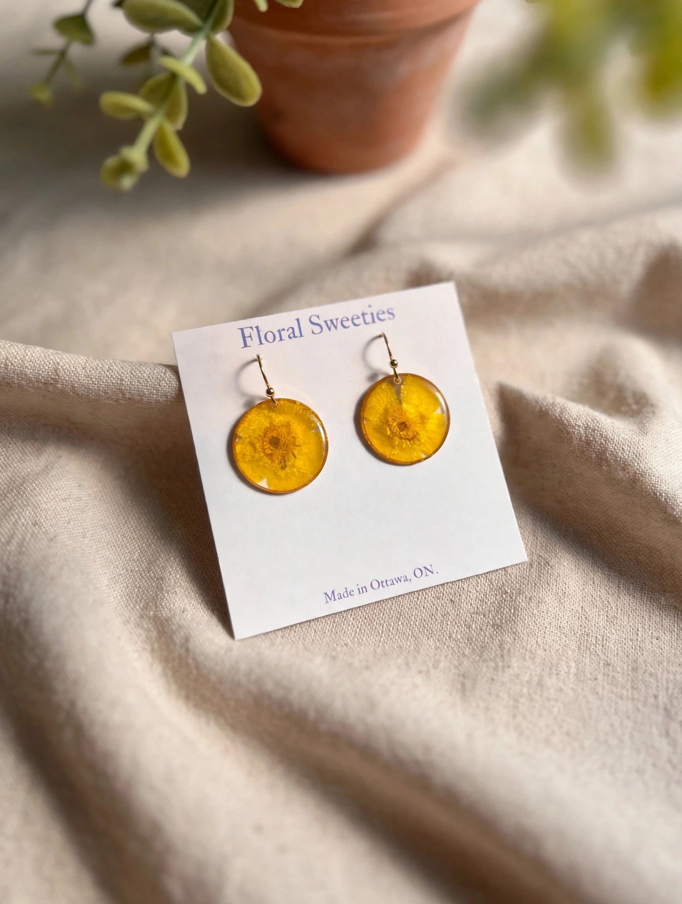 Buttercup flower dangle earrings on a display card on draped linen with a terracotta pot in the background