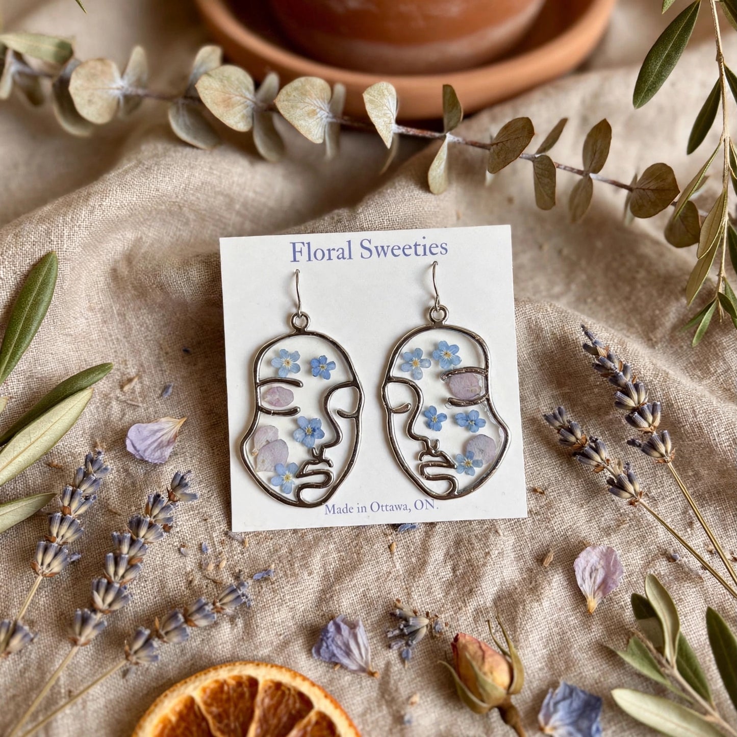 Abstract face dangle earrings with forget-me-not and hydrangea flowers in resin, styled on linen