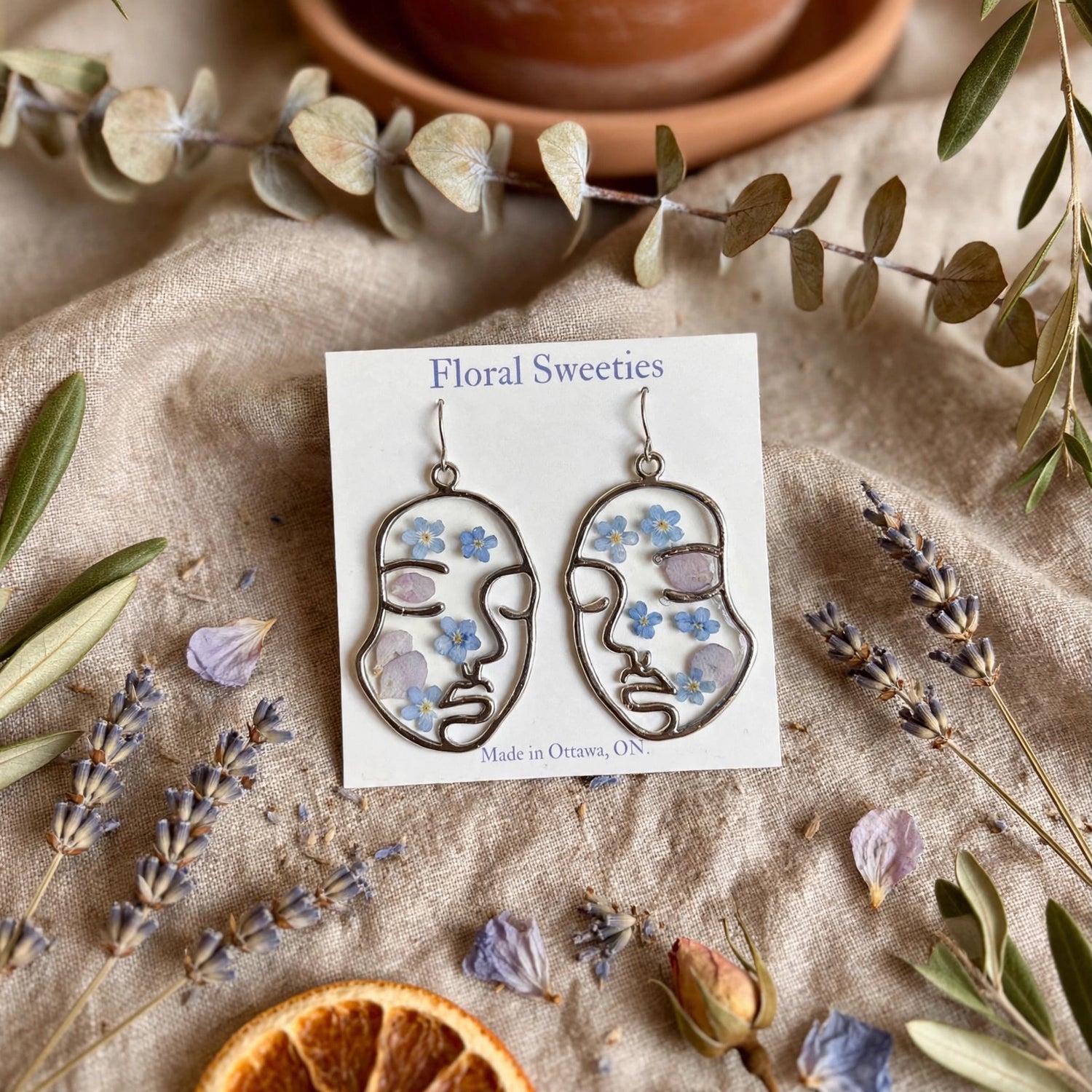 Abstract face dangle earrings with forget-me-not and hydrangea flowers in resin, styled on linen