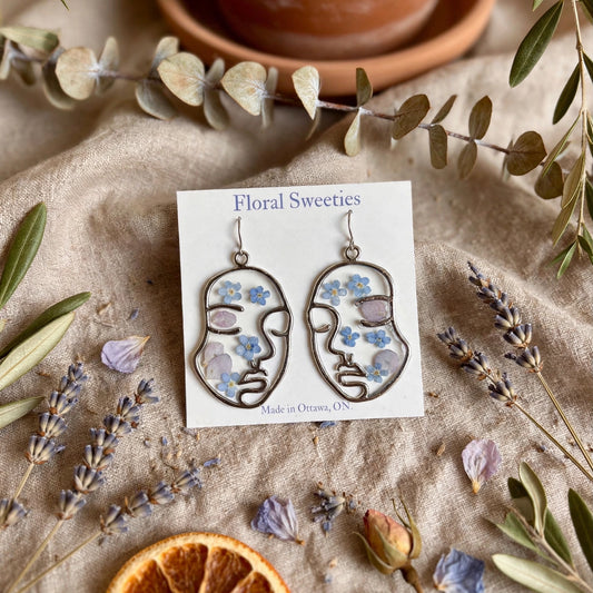 Abstract face dangle earrings with forget-me-not and hydrangea flowers in resin, styled on linen