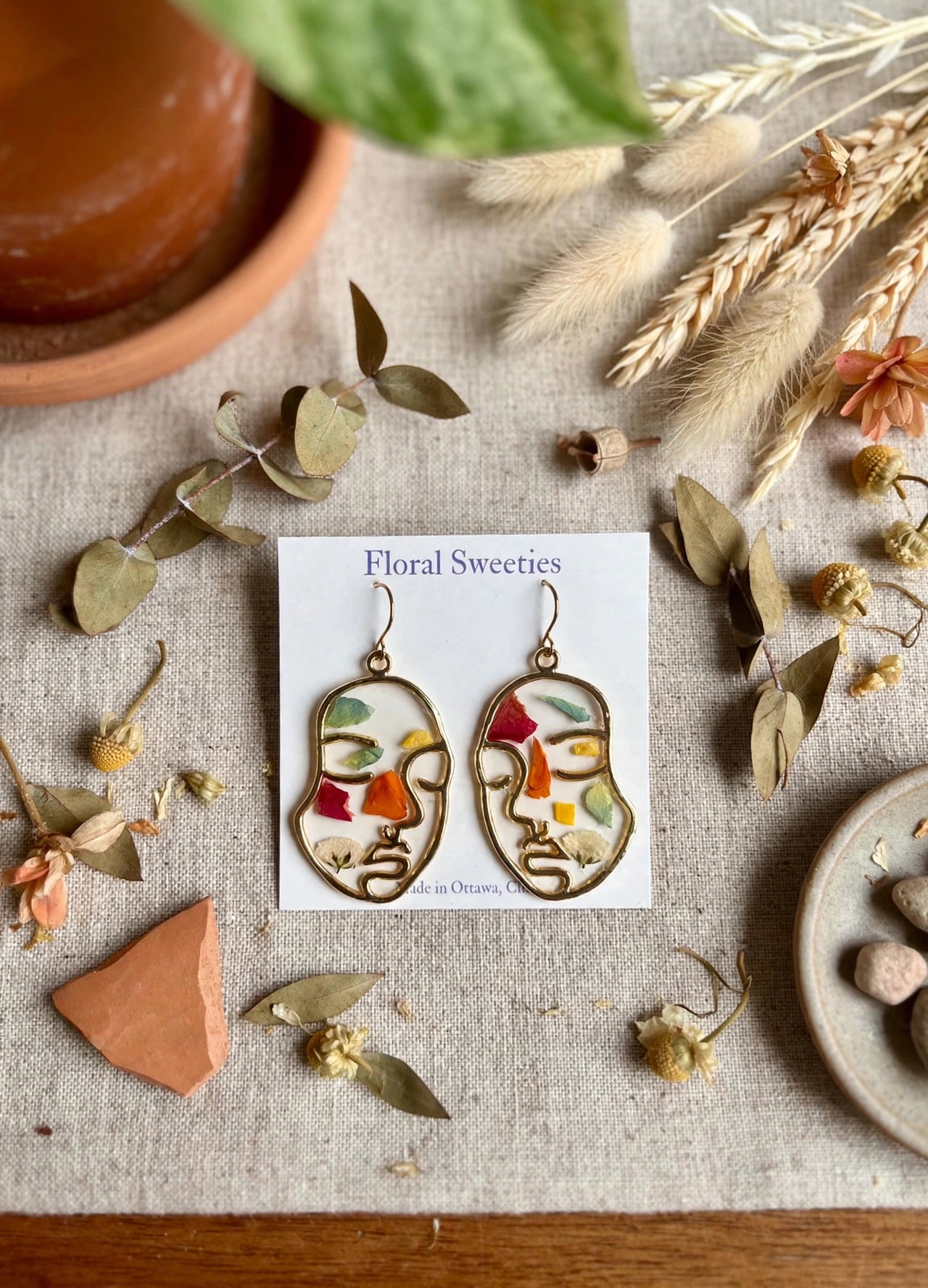 Abstract face dangle earrings with mixed pressed flower petals in resin, styled on linen
