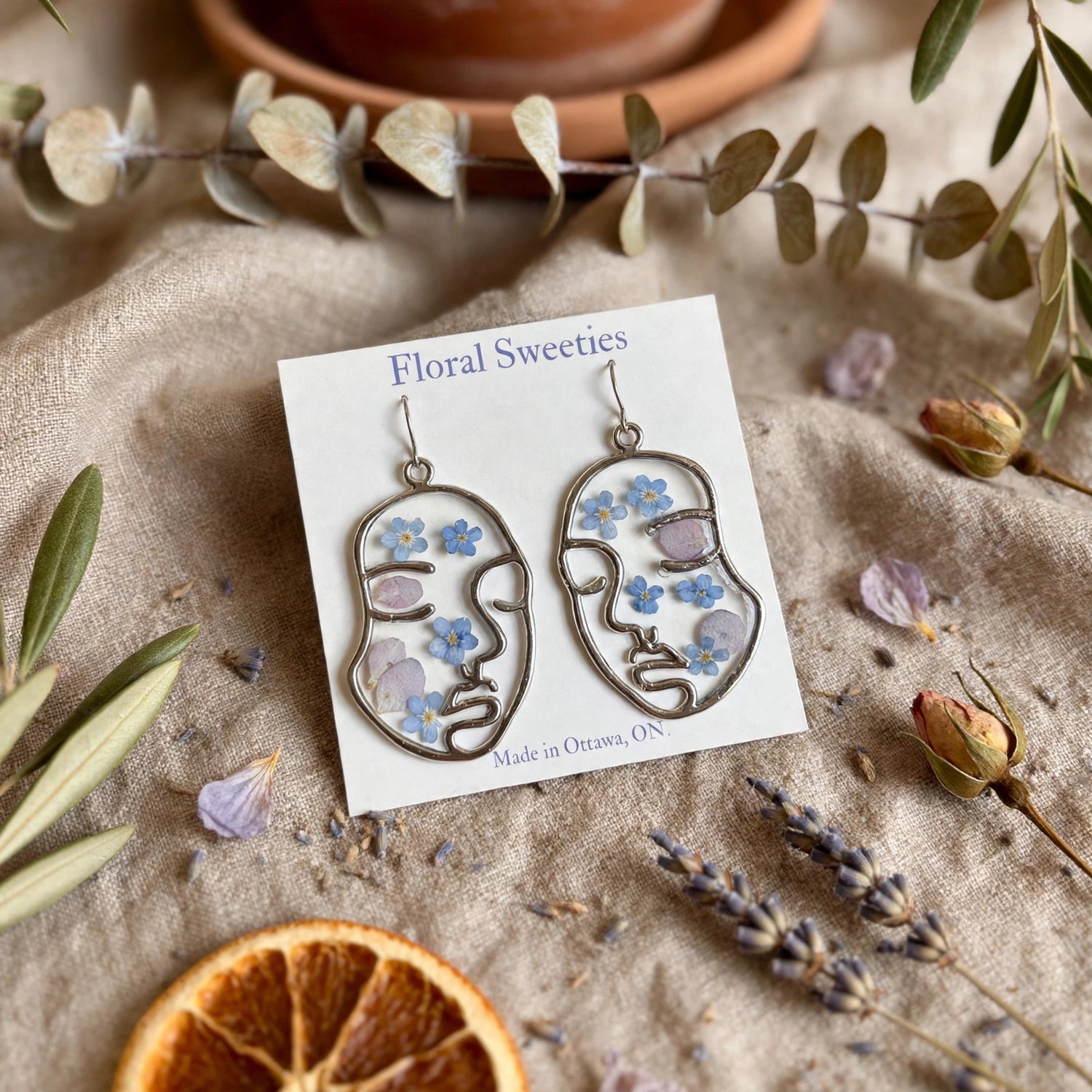 Abstract face earrings with forget-me-nots and hydrangea, angled view with dried orange slice
