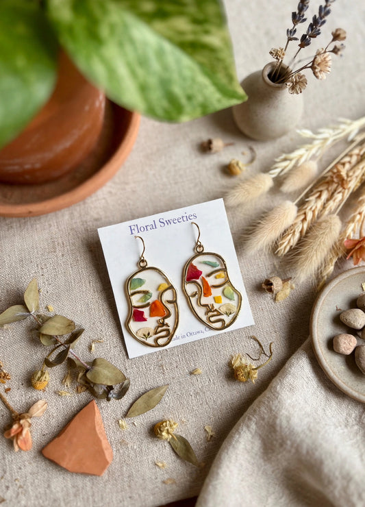 Abstract face earrings with mixed pressed petals, angled view with dried grasses