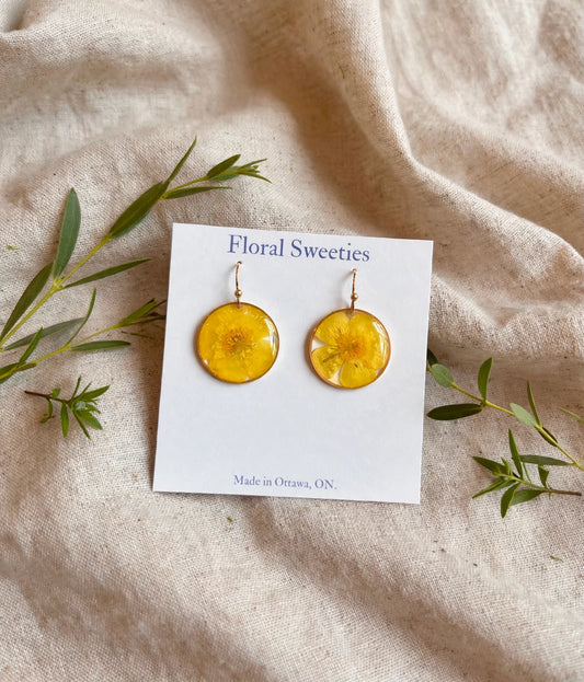 Angled view of buttercup flower resin dangle earrings on a display card on draped linen with greenery