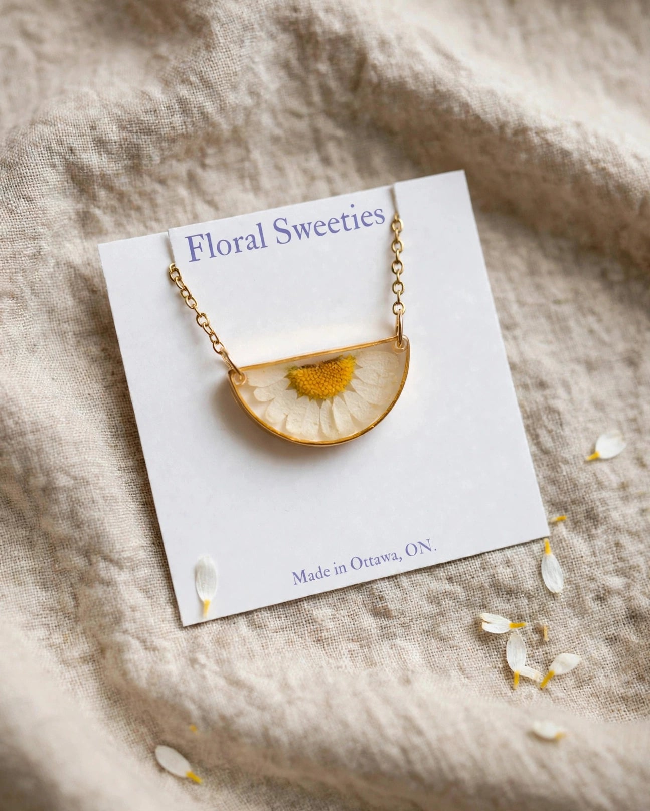 Close-up angled view of the daisy resin pendant necklace on its display card on linen with loose daisy petals