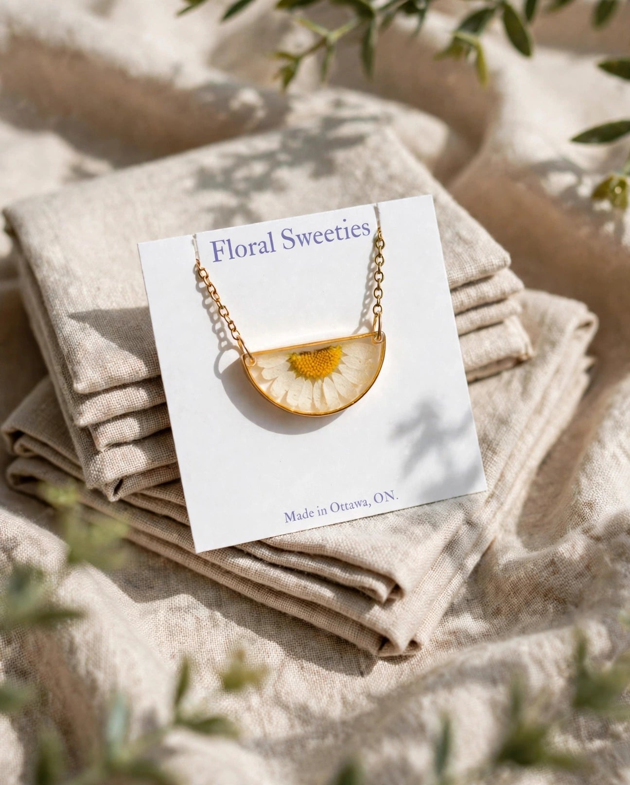 Daisy flower necklace on its display card resting on draped linen with greenery shadows