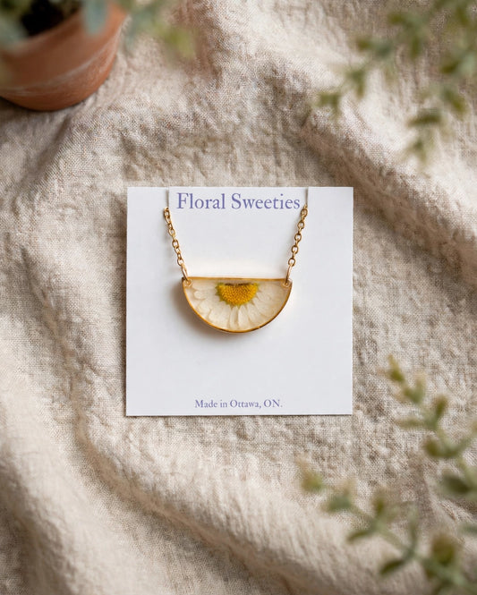 Daisy flower necklace with a real pressed daisy in a gold half-circle resin pendant on a display card, styled on linen