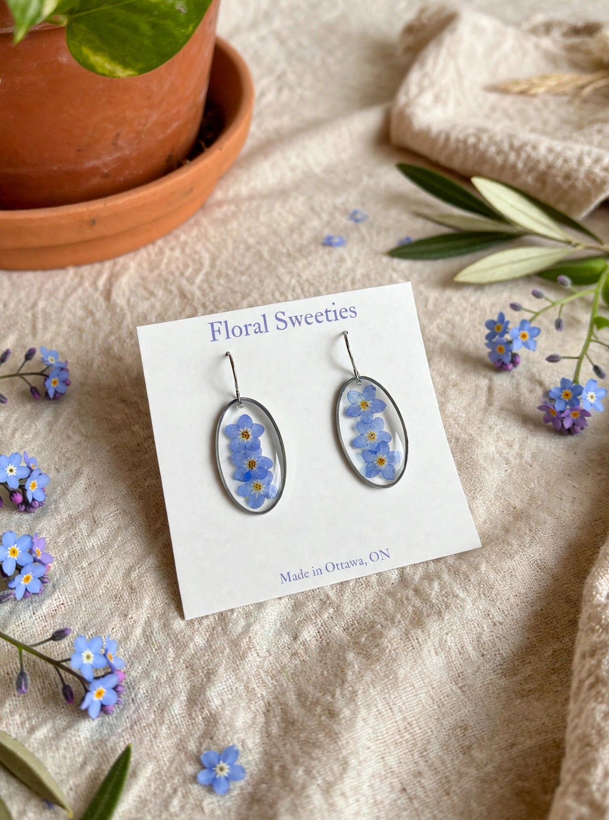 Forget-me-not oval dangle earrings angled view with fresh forget-me-not sprigs on linen