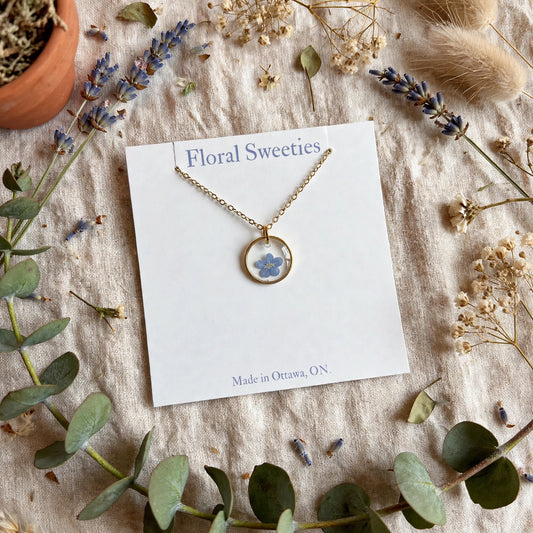 Forget-me-not necklace on its display card at an angle, with lavender and eucalyptus on linen