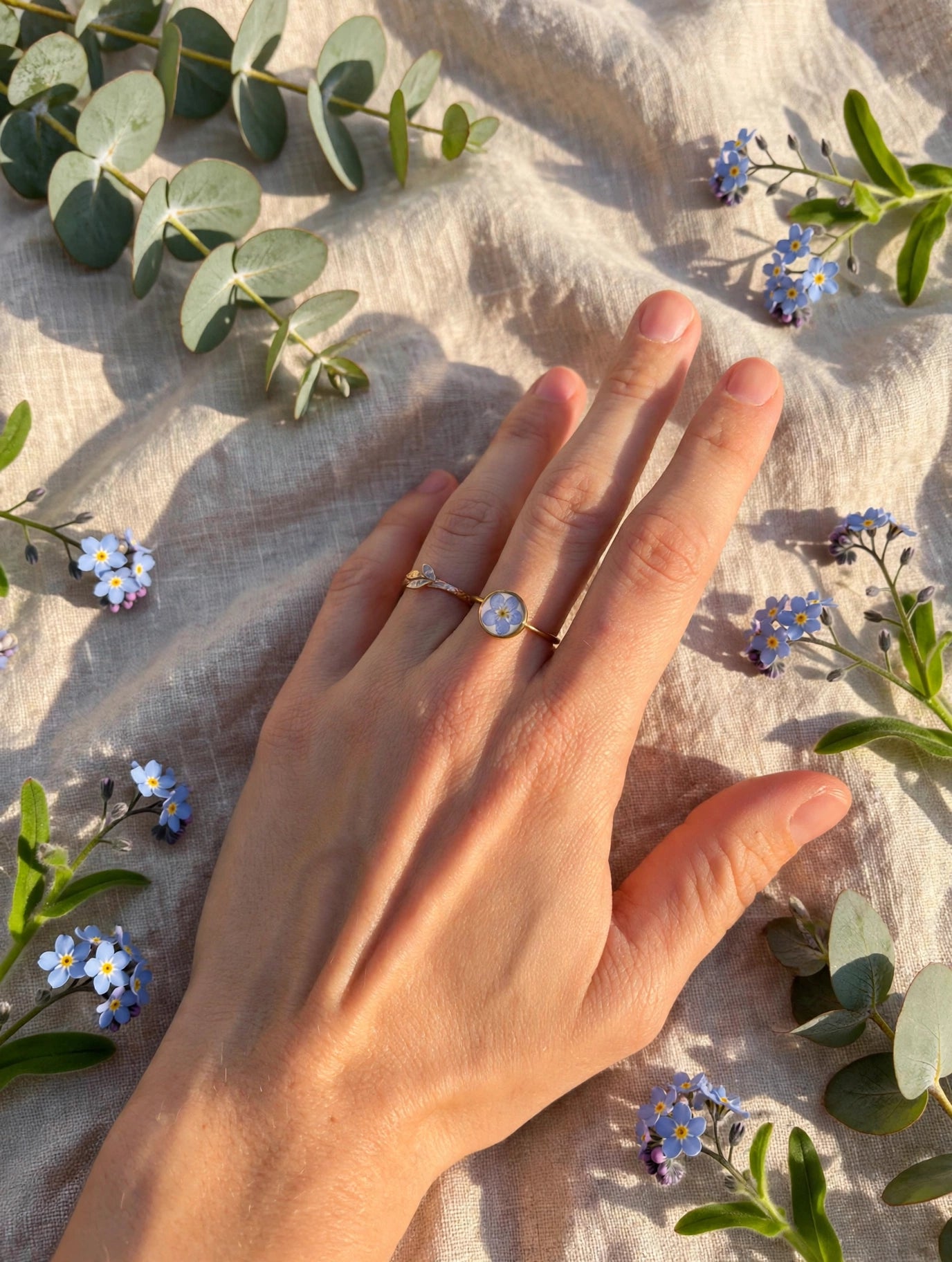 Gold forget-me-not ring worn on a hand resting on linen with forget-me-not sprigs