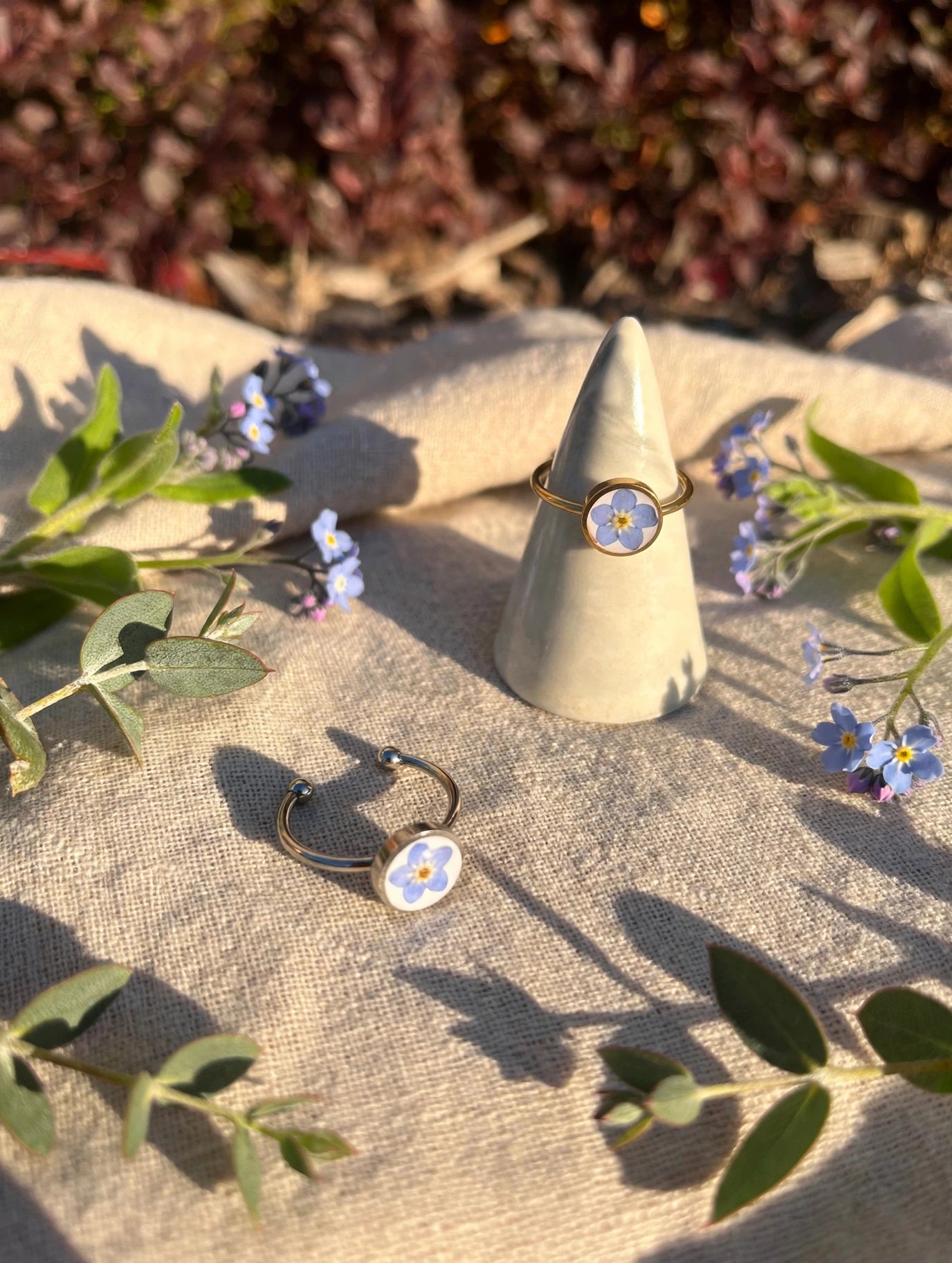 Silver forget-me-not ring with open adjustable band beside the gold ring on a cone, sunlight on linen